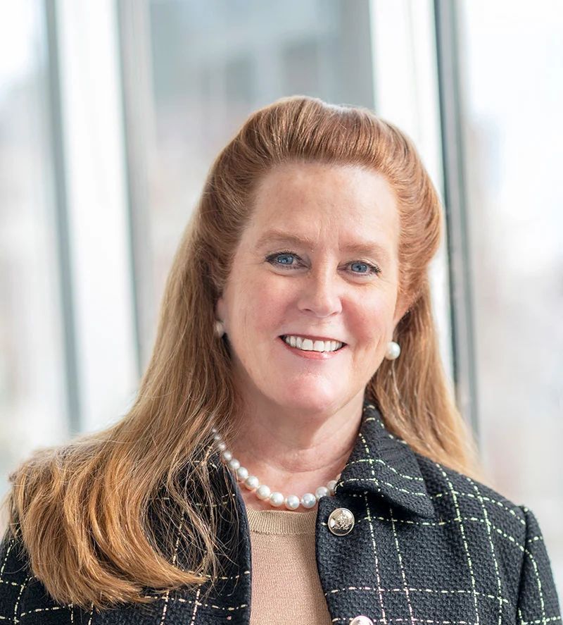 A woman wearing a black jacket and a pearl necklace is smiling for the camera.