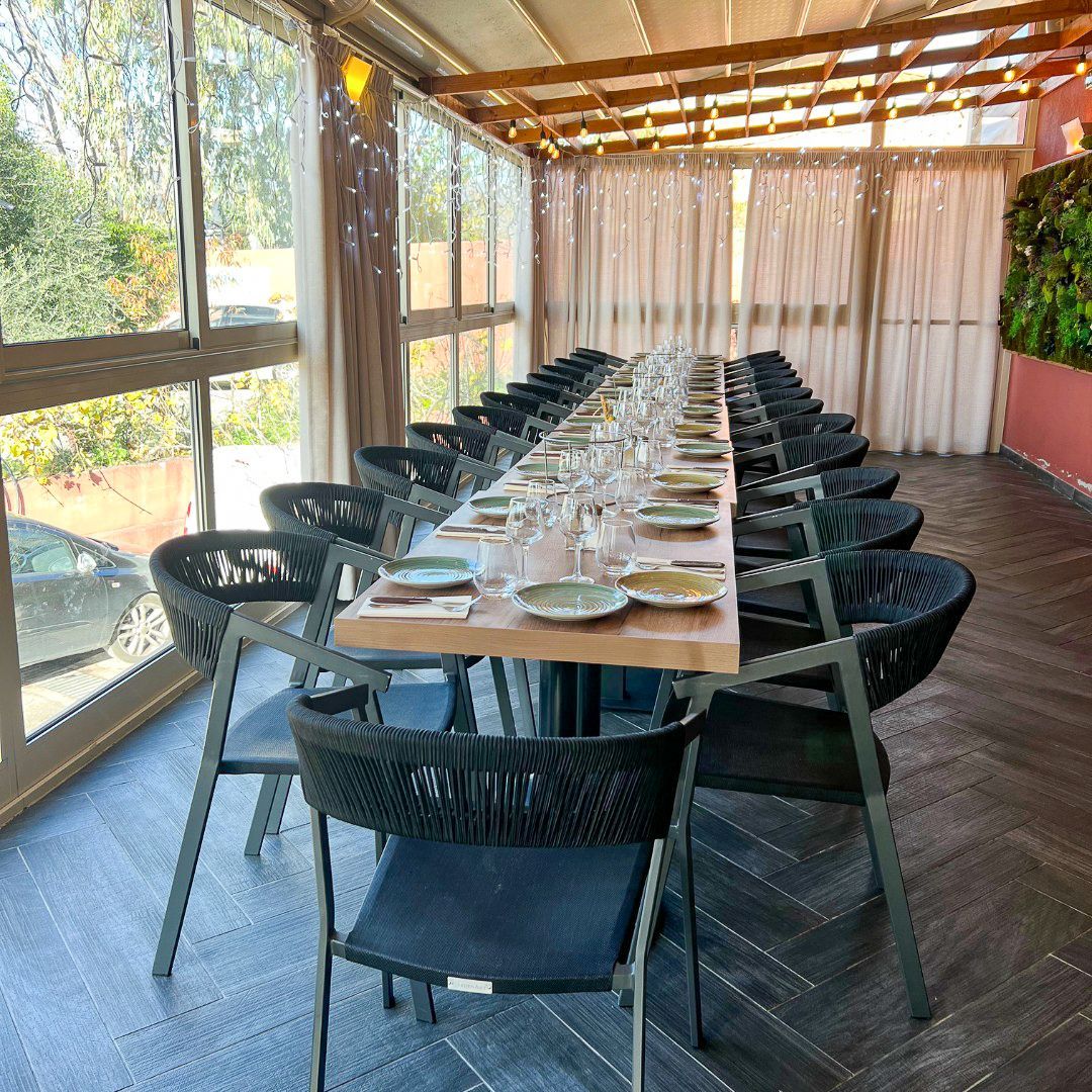 Longue table en bois dressée pour un repas dans une véranda avec chaises, fenêtres et verdure décorative.