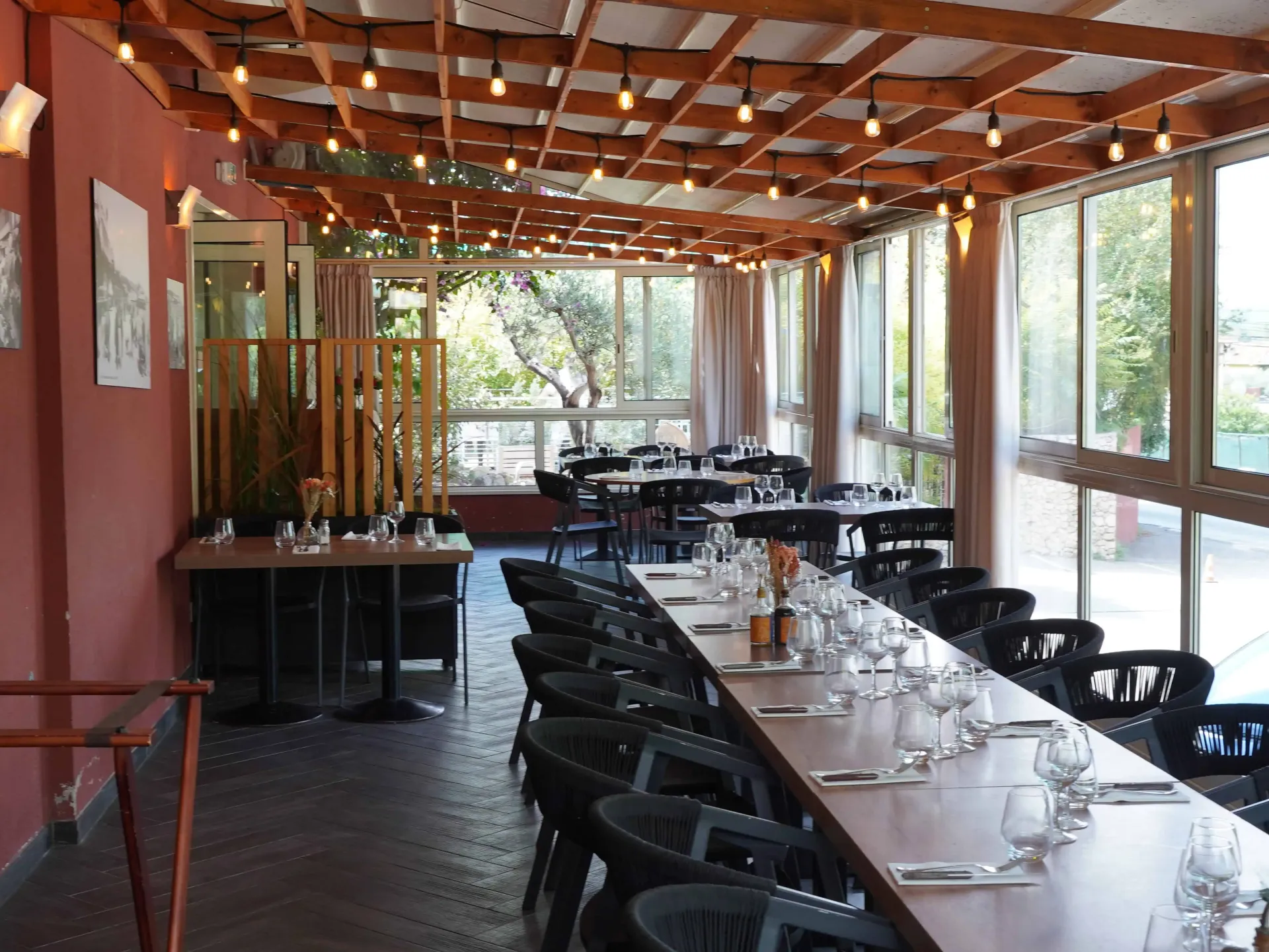 Intérieur de restaurant avec longues tables, chaises noires et guirlandes lumineuses, entouré de grandes fenêtres.