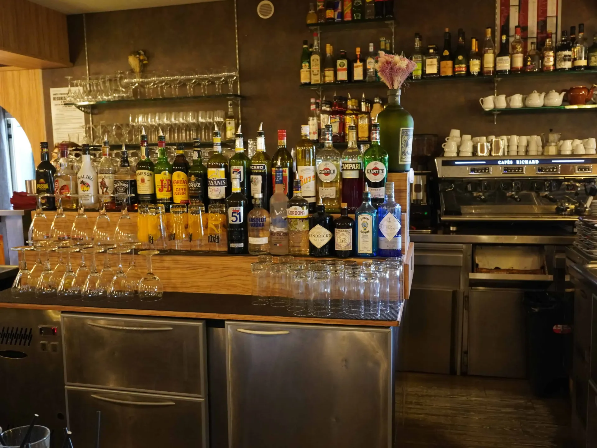 Bar avec bouteilles, verres et machine à expresso. Des étagères accueillent boissons et verres sur le comptoir.