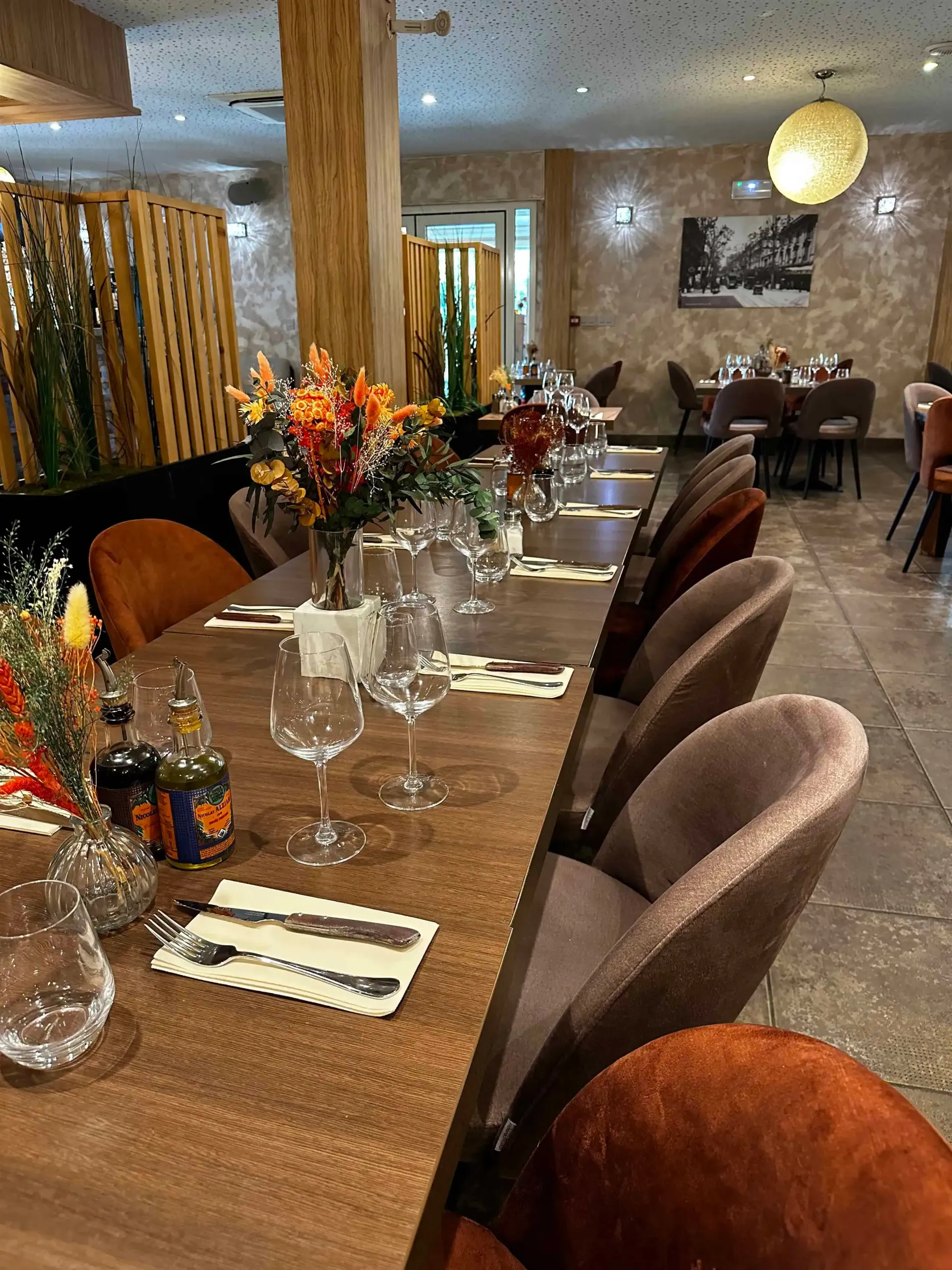 Longue table en bois dressée pour les convives d'un restaurant avec verres à vin, fleurs et chaises.