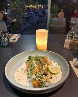 Une assiette de poisson pané avec une sauce crémeuse, un quartier de citron vert et de la verdure, servie dans un restaurant en plein air à la lueur des bougies.