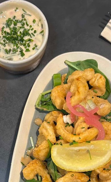 Une assiette de calamars frits avec des légumes verts, des oignons marinés et un quartier de citron, accompagnée d'un petit bol de sauce crémeuse.