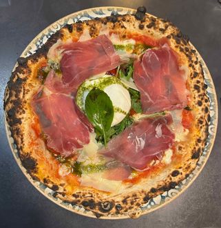 Une pizza napolitaine avec sauce tomate, tranches de prosciutto, pesto, copeaux de fromage et une feuille de basilic frais sur le dessus.