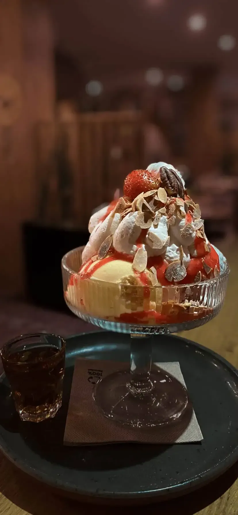 Coupe glacée avec plusieurs garnitures dans un bol en verre, accompagnée d'un verre de liquide foncé sur une assiette.