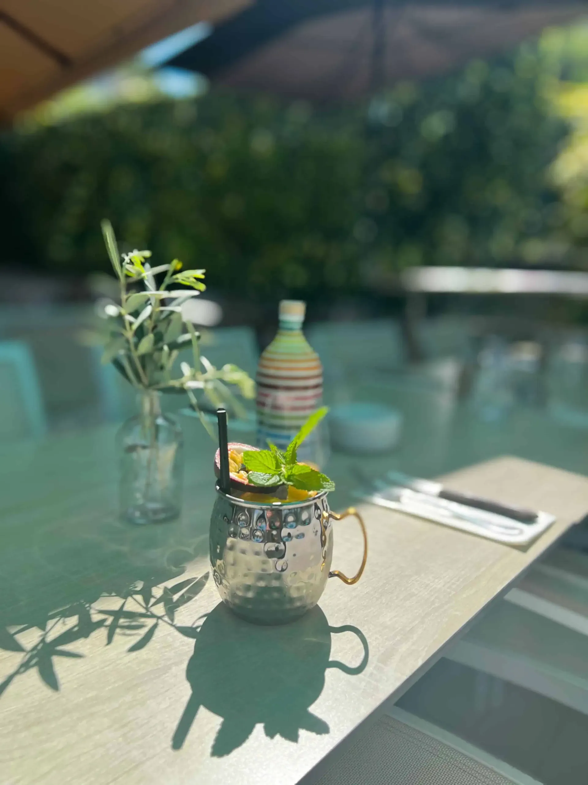 Cocktail dans une tasse en argent sur une table, avec des branches d'olivier et une bouteille décorative en arrière-plan.