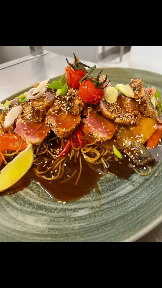 Thon poêlé et légumes sur des nouilles dans une assiette verte, garni de tomates et de graines de sésame.