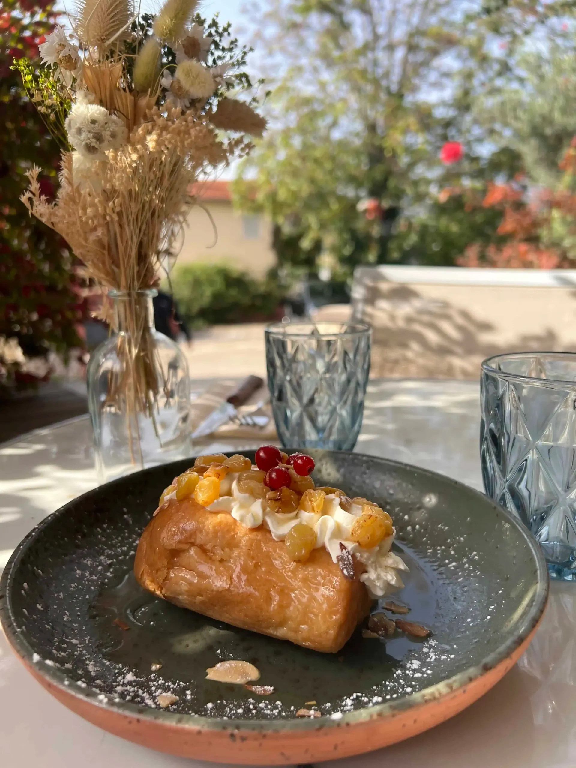 Une pâtisserie à la crème et aux fruits sur une assiette, avec deux verres bleus et un vase de fleurs séchées dehors.