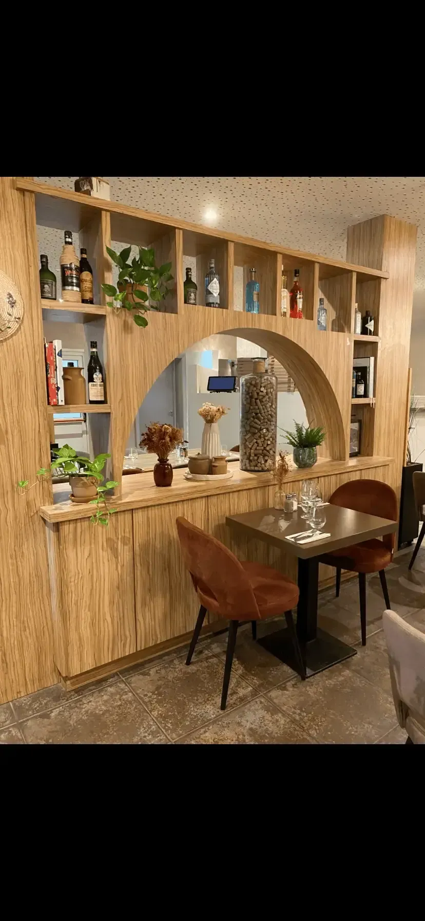Intérieur de restaurant avec étagère en bois arquée, bouteilles, plantes, petite table et chaises.