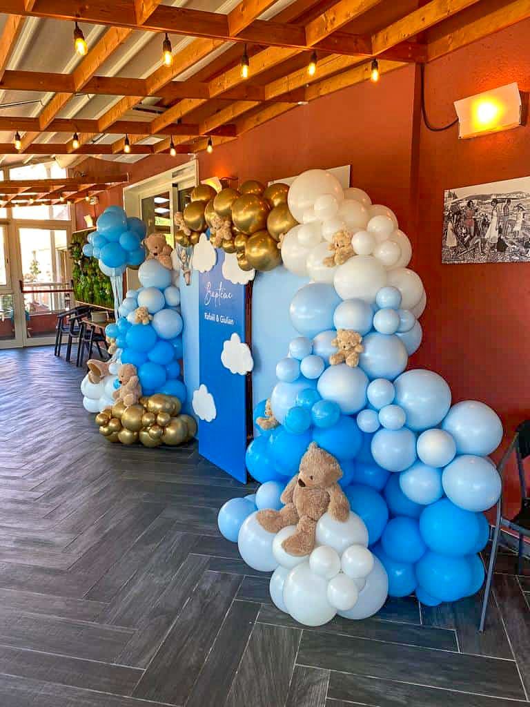 Une arche de ballons bleus, blancs et dorés décore une porte de baby shower, avec ours en peluche et découpes de nuages.