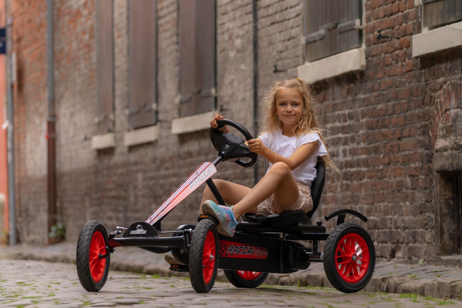 Ein Mädchen sitzt fröhlich auf einem roten Speedy Go-Kart von Dino Cars in einer gepflasterten Gasse.