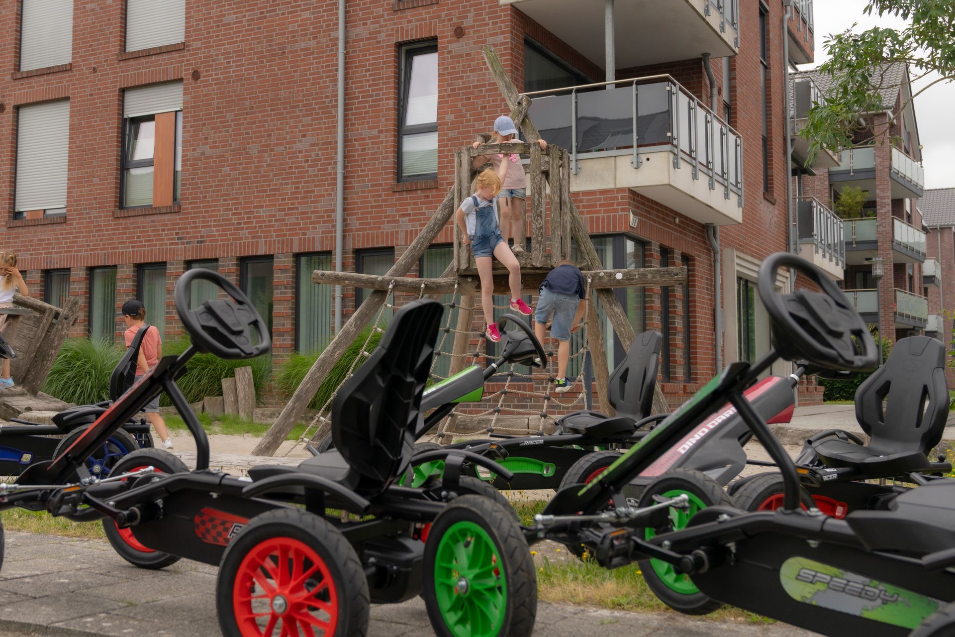 Children playing on a wooden structure near pedal go-karts in front of a brick building.