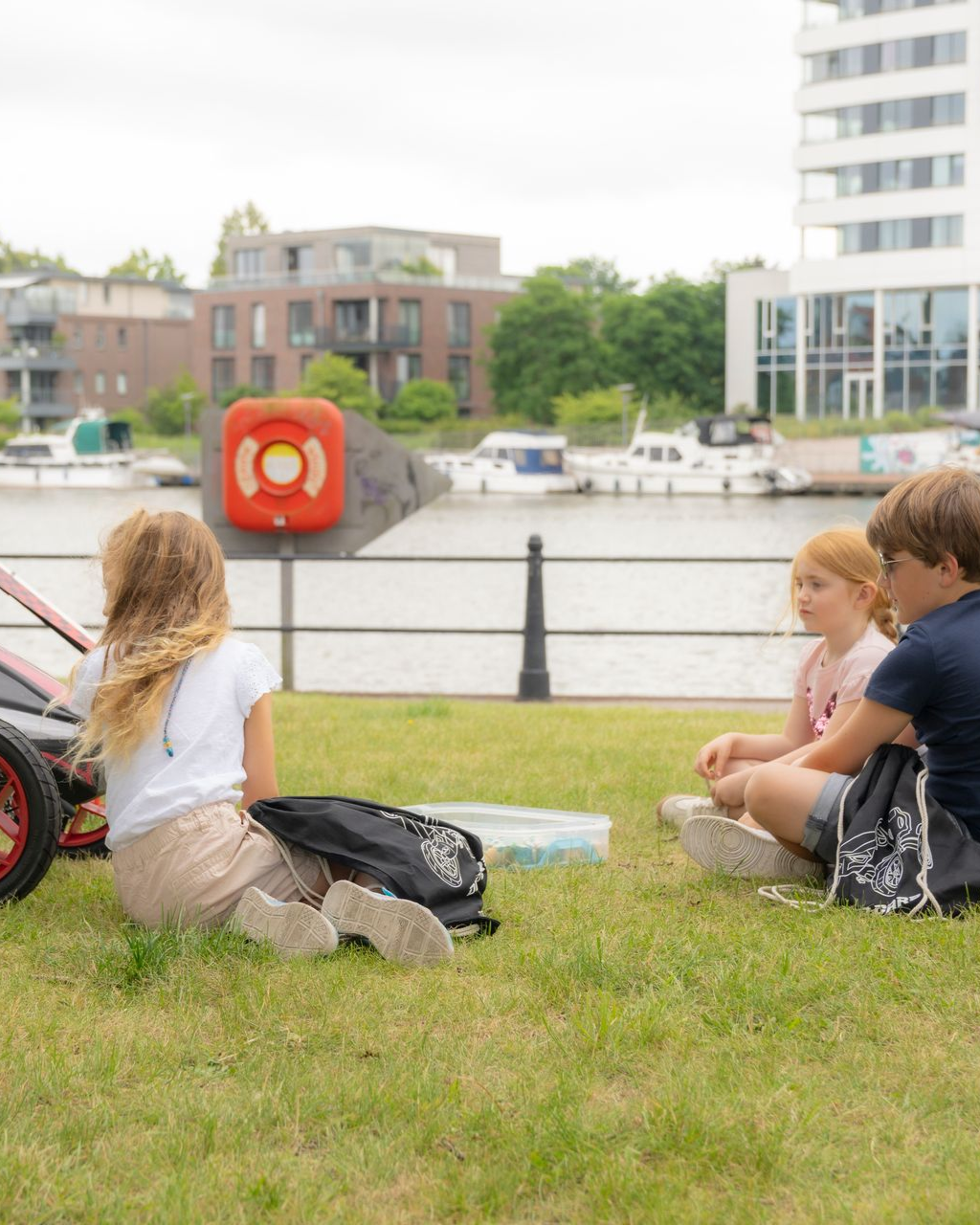 Drei Kinder sitzen auf einer Wiese am Hafen und unterhalten sich, links im Bild ist ein Dino Cars Racer Go-Kart zu sehen.