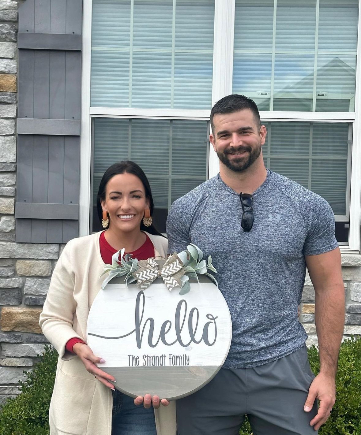 A man and a woman are standing in front of a house holding a hello sign.