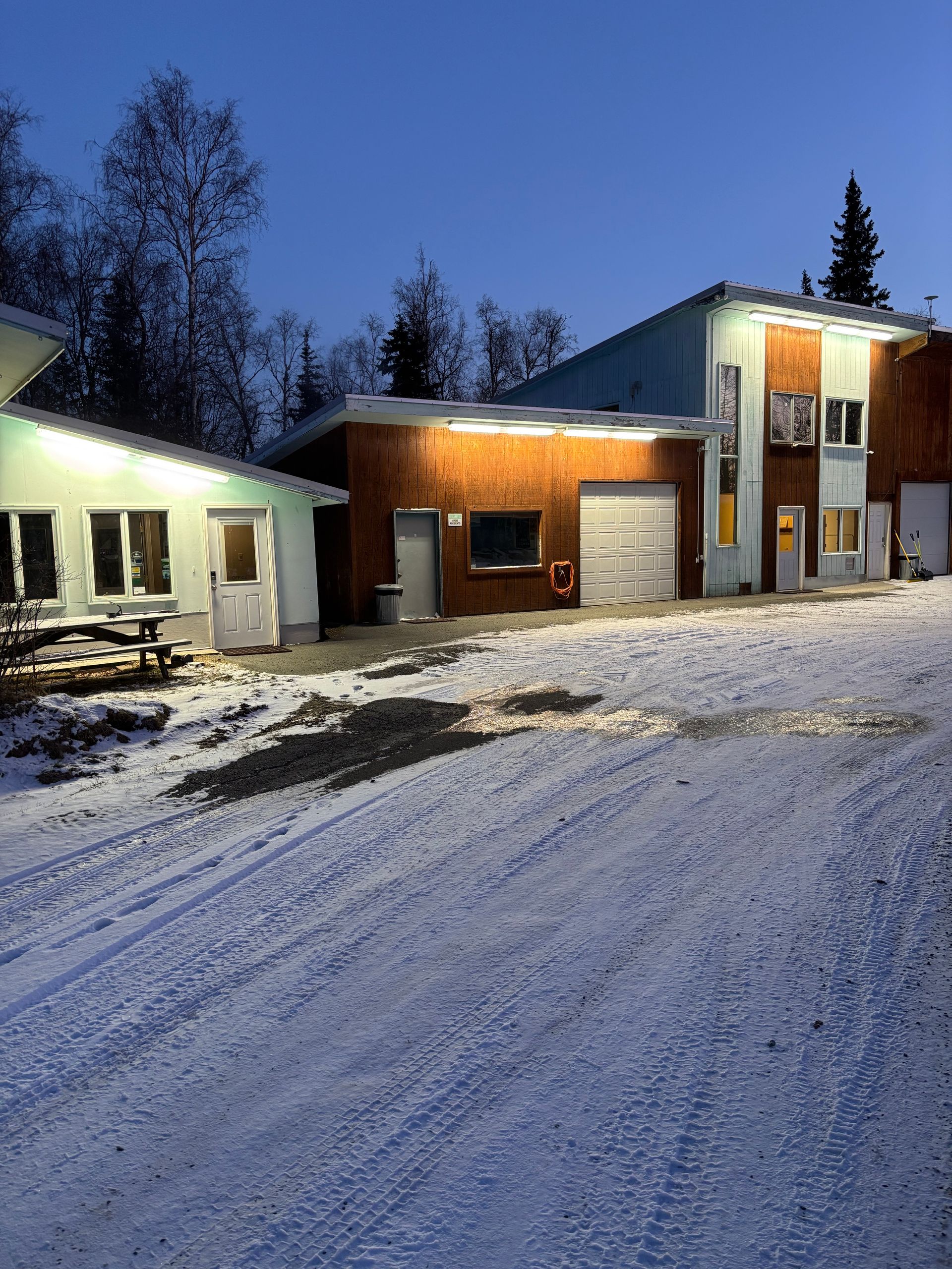 Buildings in a snowy lot at dusk, with trees in the background. One has a garage door.