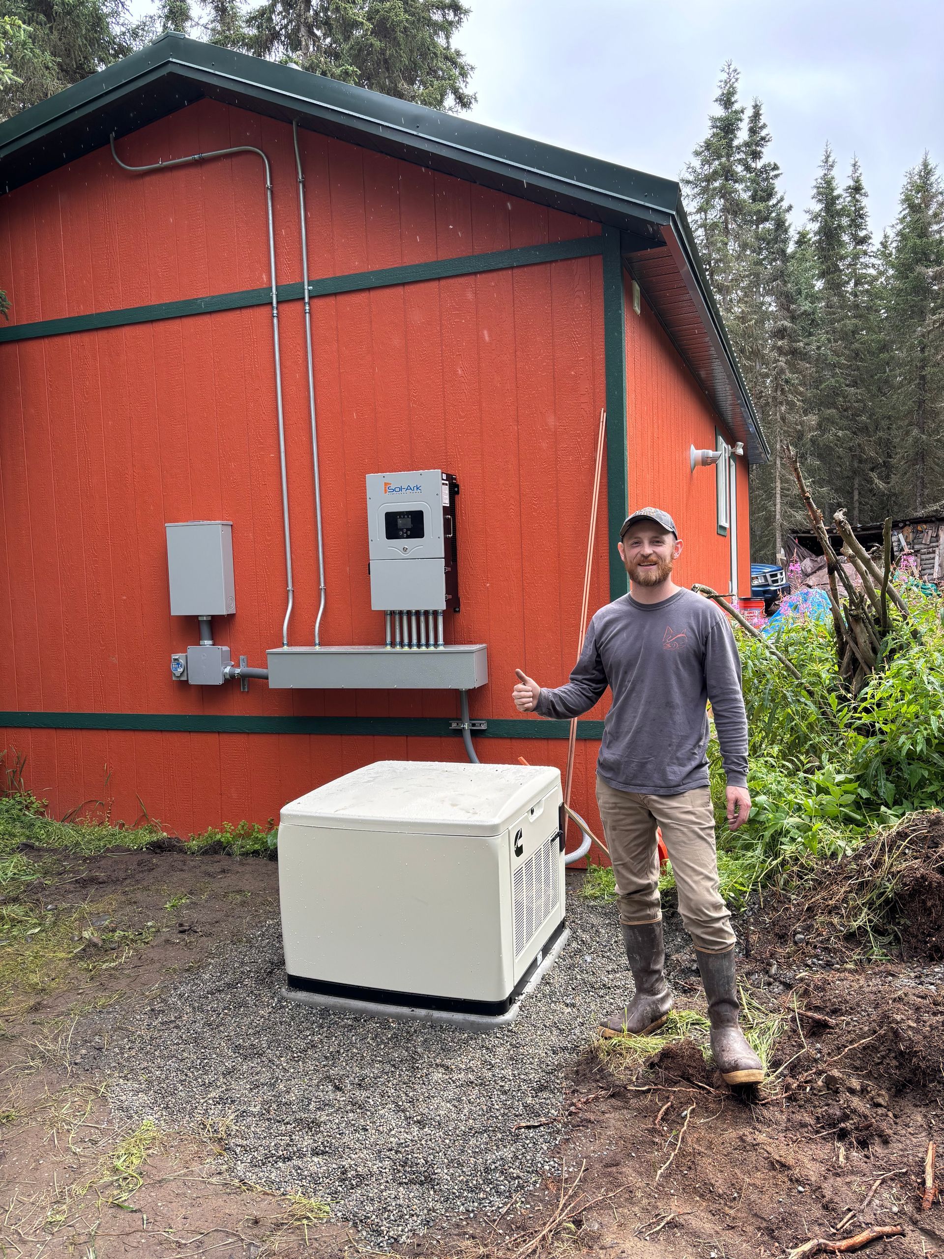 Man standing next to a building with solar equipment and a generator. Orange building, cloudy sky, gravel ground.