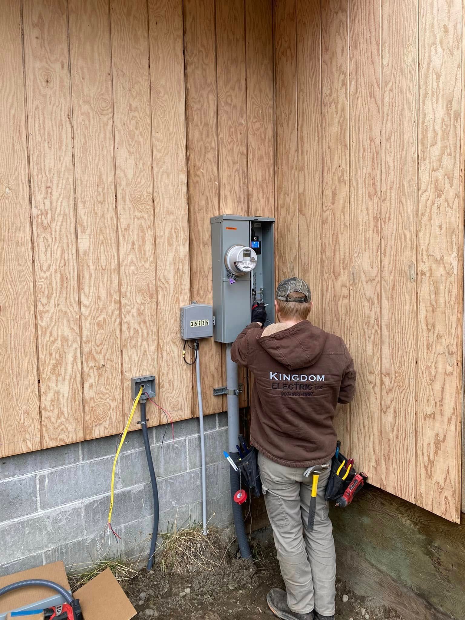 Electrician working on electrical panel mounted on plywood siding of a building.