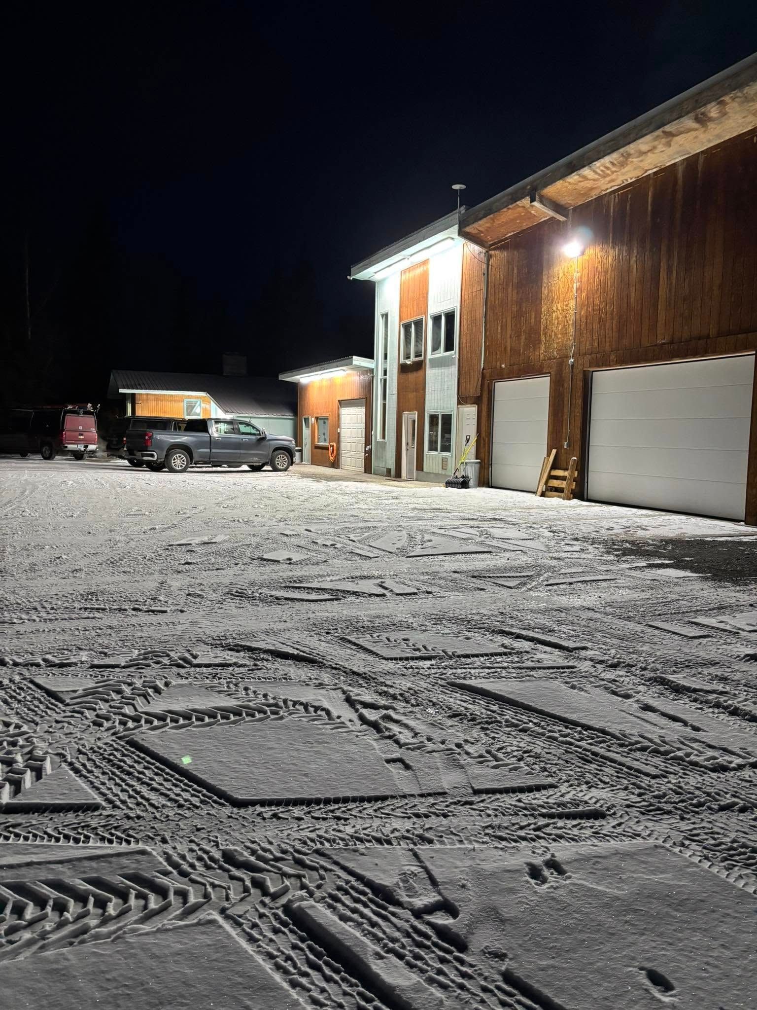 Snow-covered parking area at night; truck parked near a building with garage doors; tire tracks in the snow.