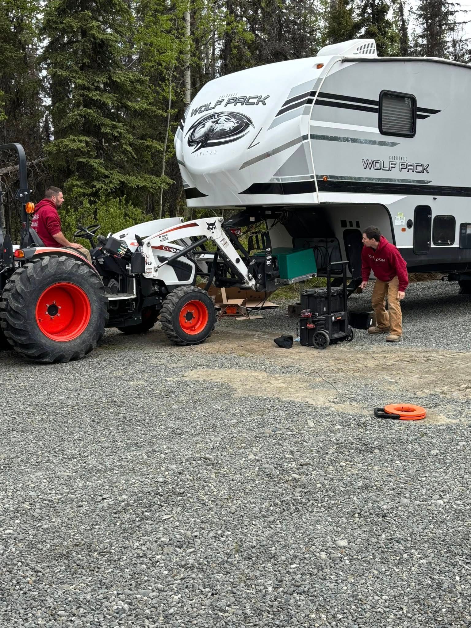 Two people using a tractor to work on an RV in a gravel area surrounded by trees.