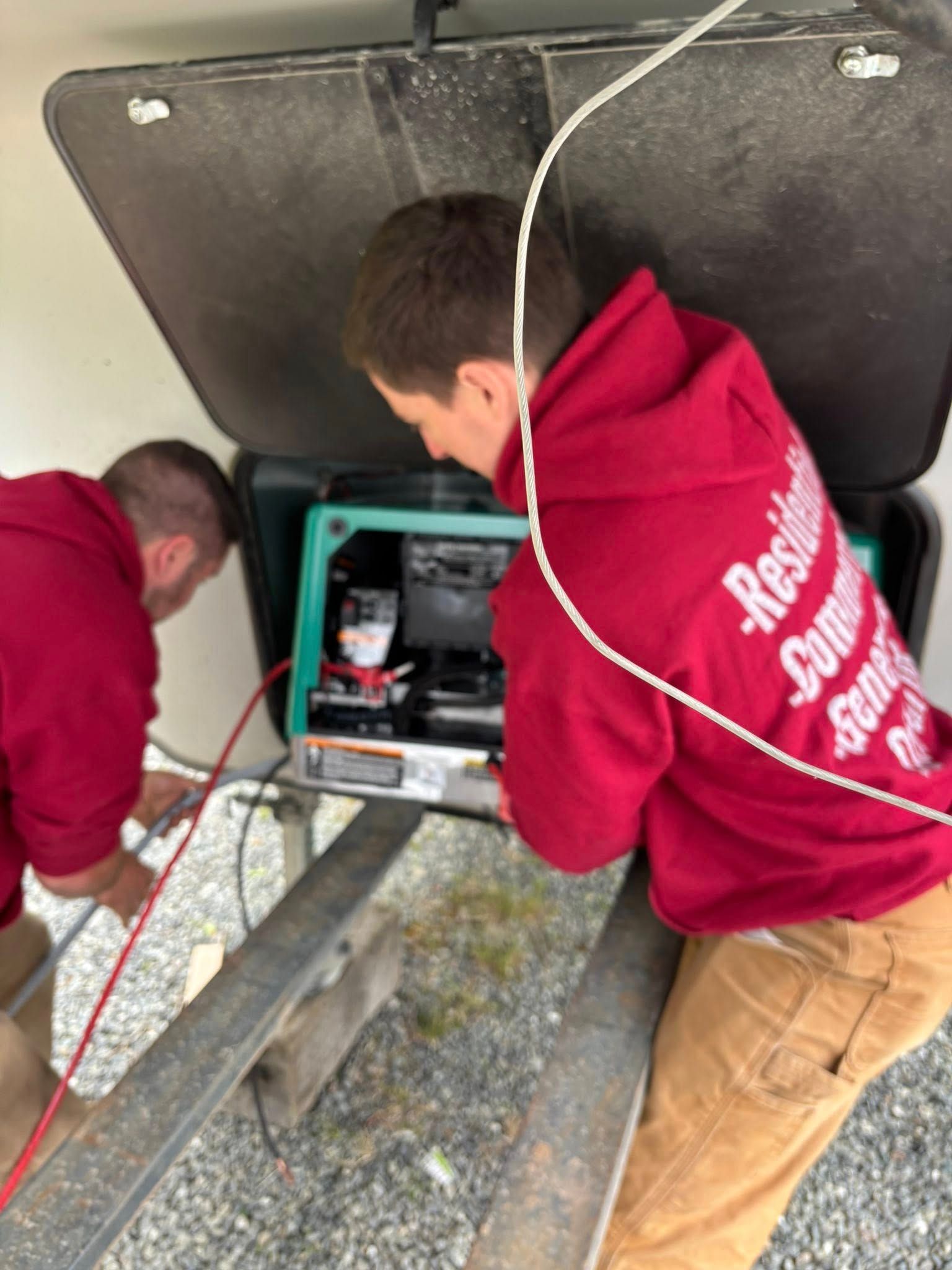 Two men in red hoodies work on a generator mounted on a trailer.