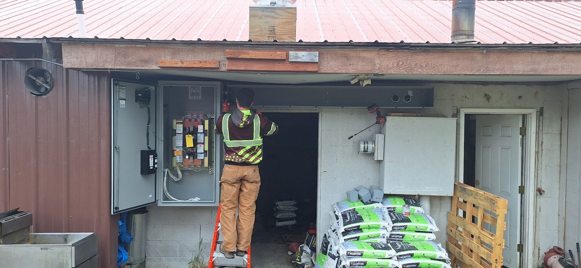 An electrician in a vest works on a panel outside a building. Bags of goods and a pallet are nearby.
