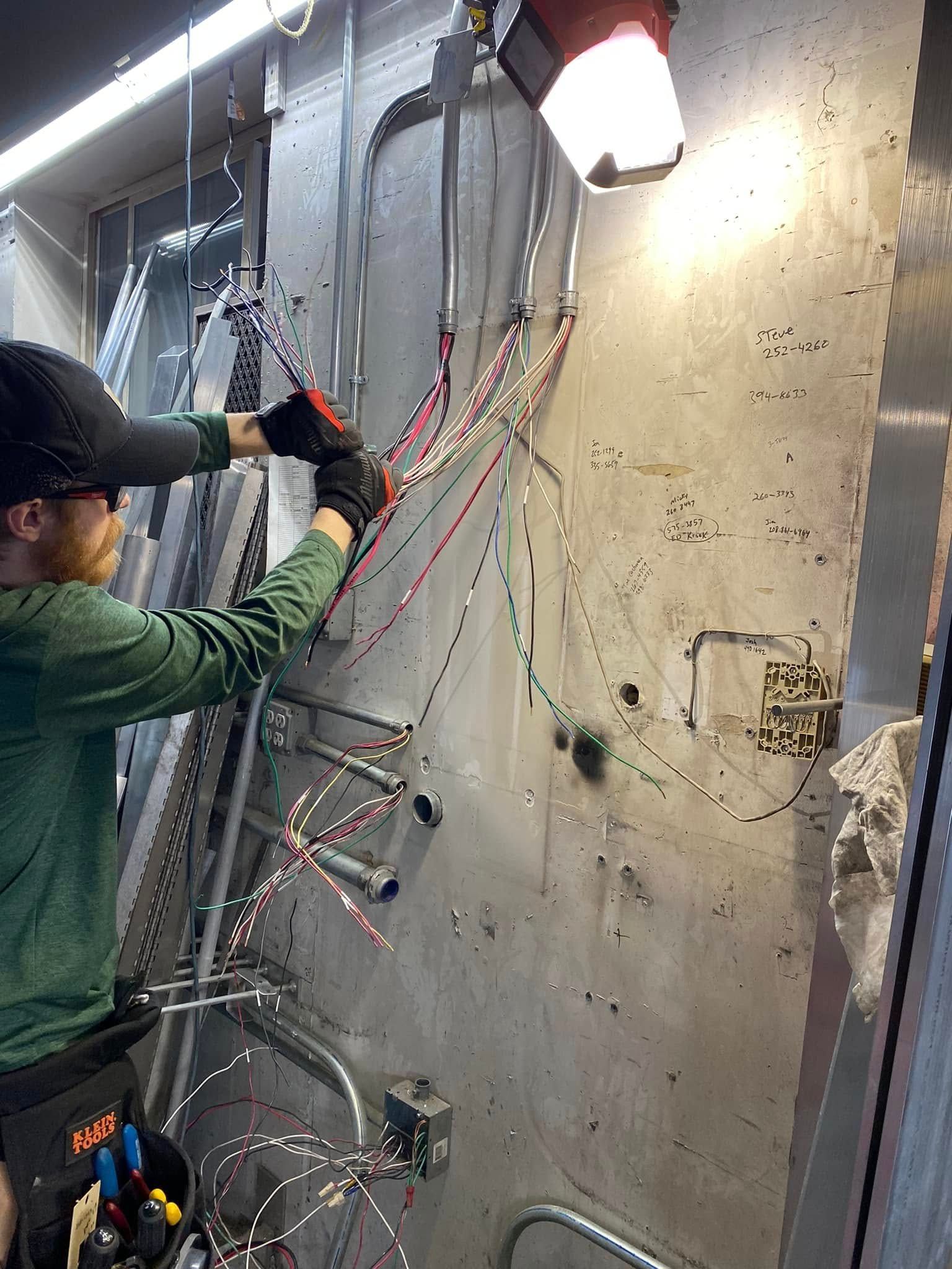 Electrician wiring cables in a metal enclosure; wearing gloves, green shirt, and hat.