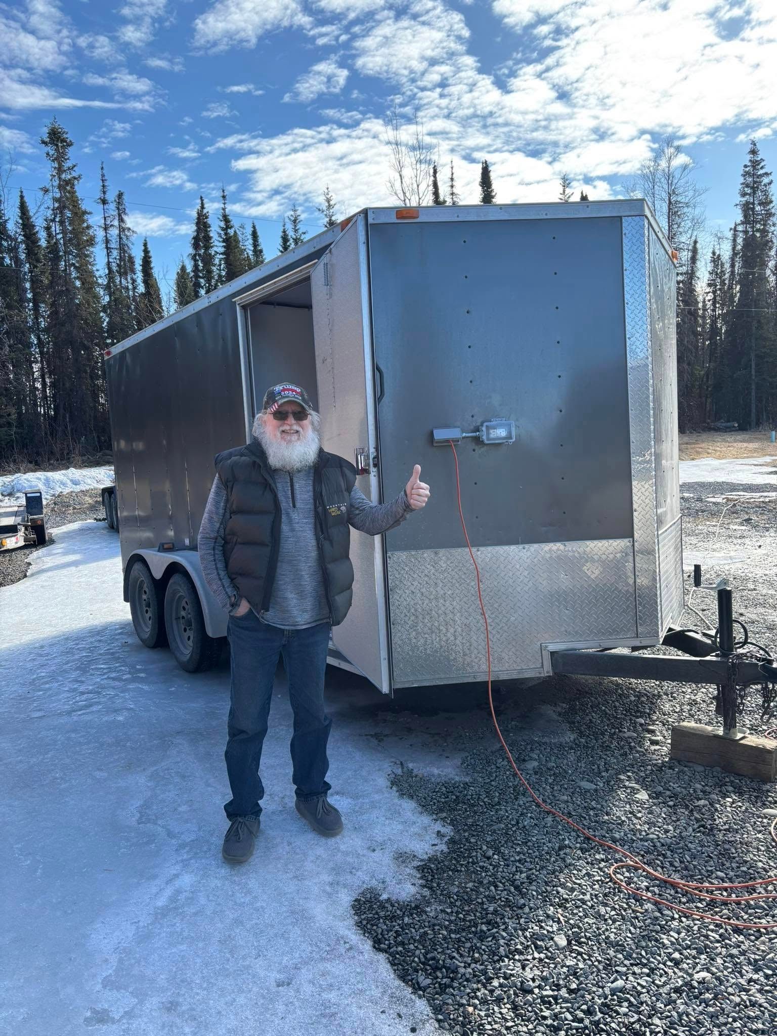 Man in front of large cargo trailer outdoors, thumbs up. Snowy ground, trees, bright sky.