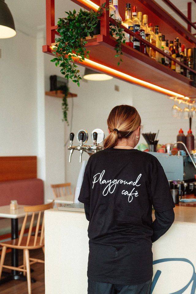 A woman wearing a black shirt that says playground cafe is standing at a counter in a restaurant.