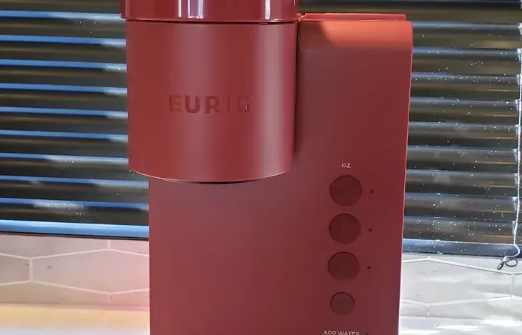 A red coffee maker is sitting on a counter next to a window.