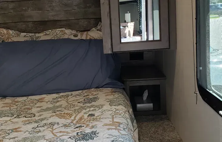 a bedroom in a rv with a bed , nightstand , mirror and window .
