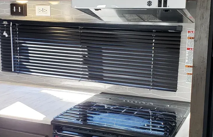 A kitchen in a rv with a stove and a window with blinds.