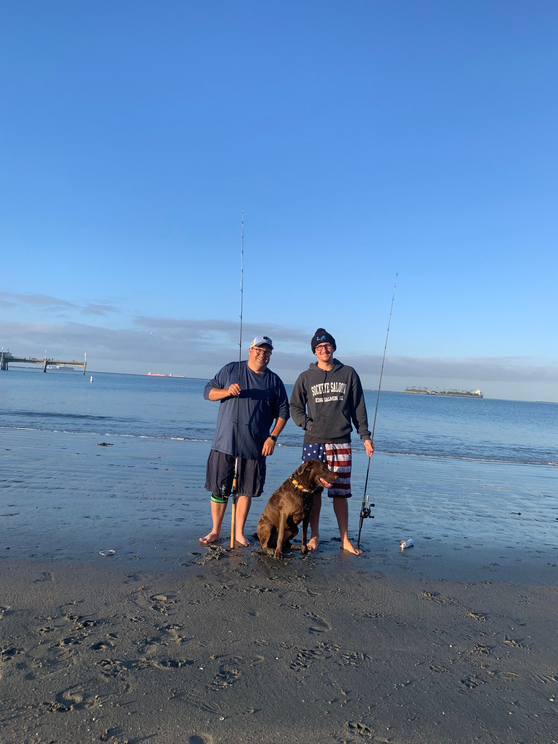 two men and a dog are standing on a beach holding fishing rods .