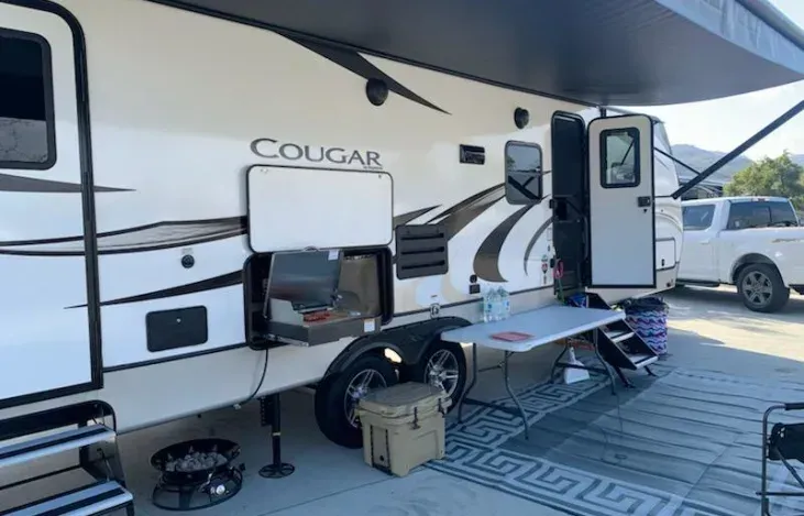 a white rv is parked in a parking lot with a table and chairs .