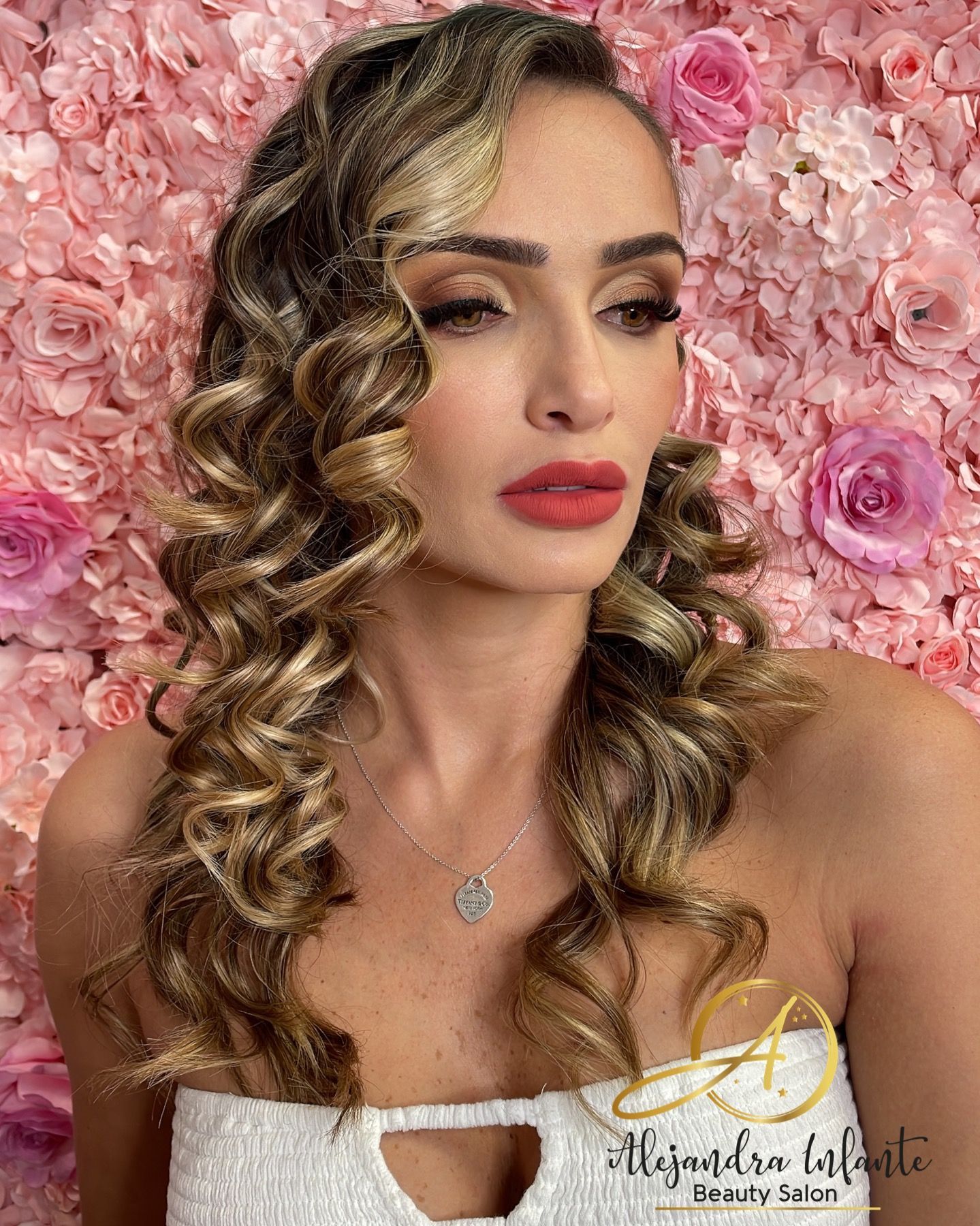 A woman with curly hair is standing in front of a wall of pink flowers.
