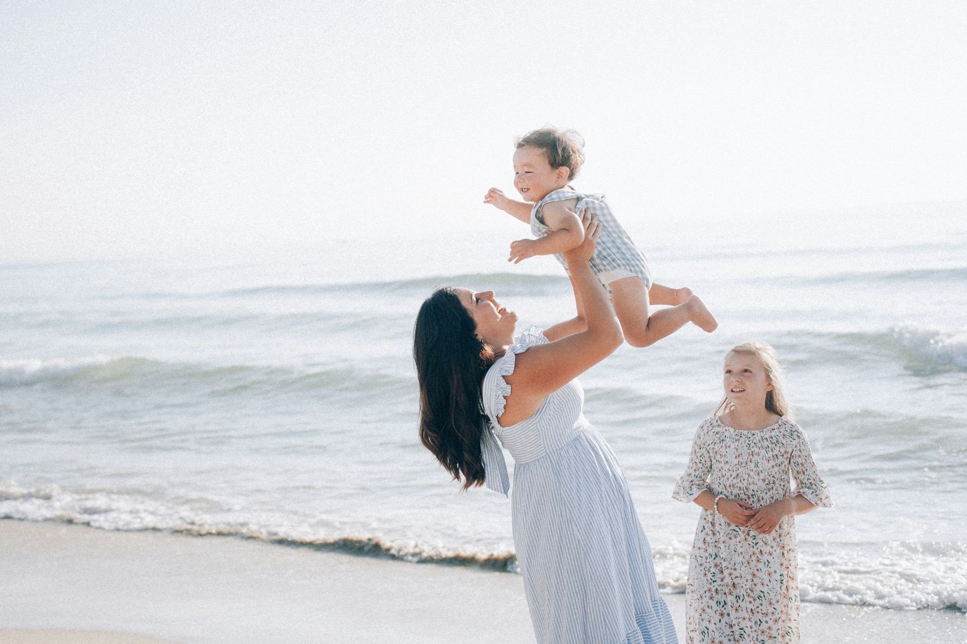 A woman is holding a baby in the air on the beach.