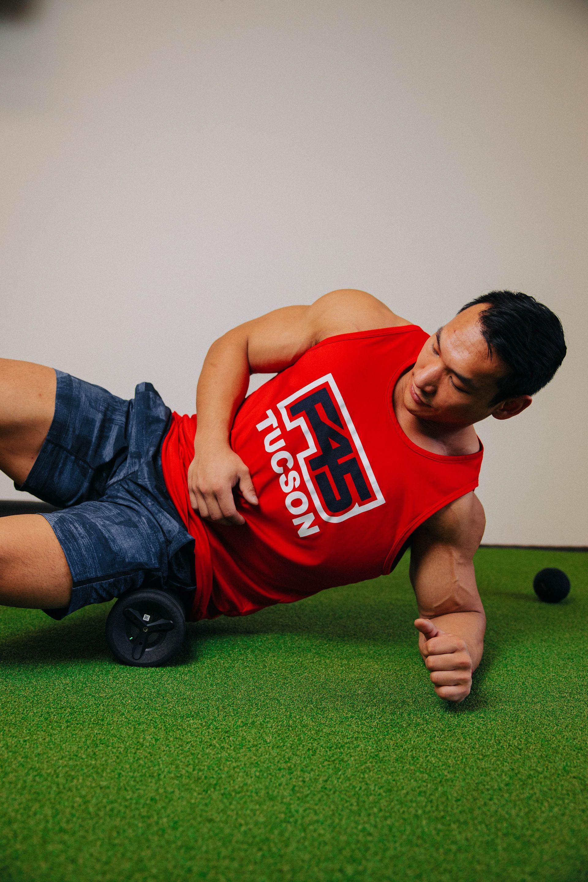 A man in a red tank top is rolling a massage roller on his leg.