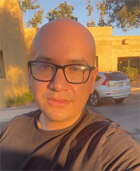 Bald person with glasses outdoors, looking at the camera. Sunlight, building, and a car in background.
