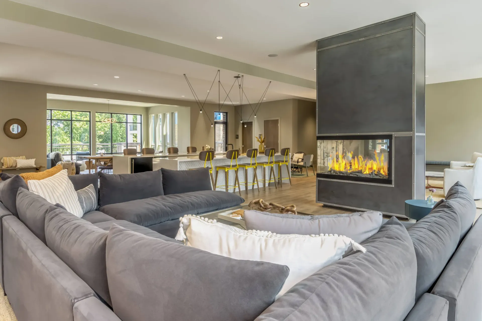 Open-concept resident lounge with a fireplace, long bar, yellow stools, and large windows.
