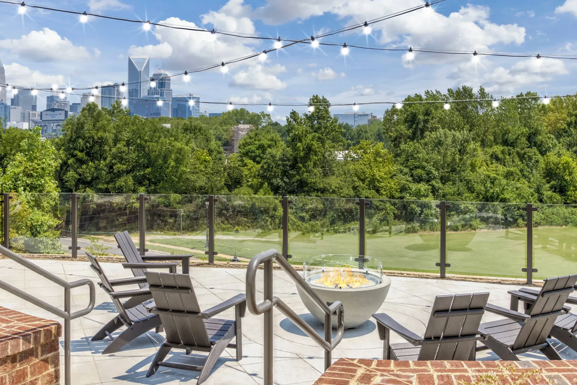 Outdoor terrace with string lights, glass railing, a fire pit, and patio chairs overlooking a city skyline.