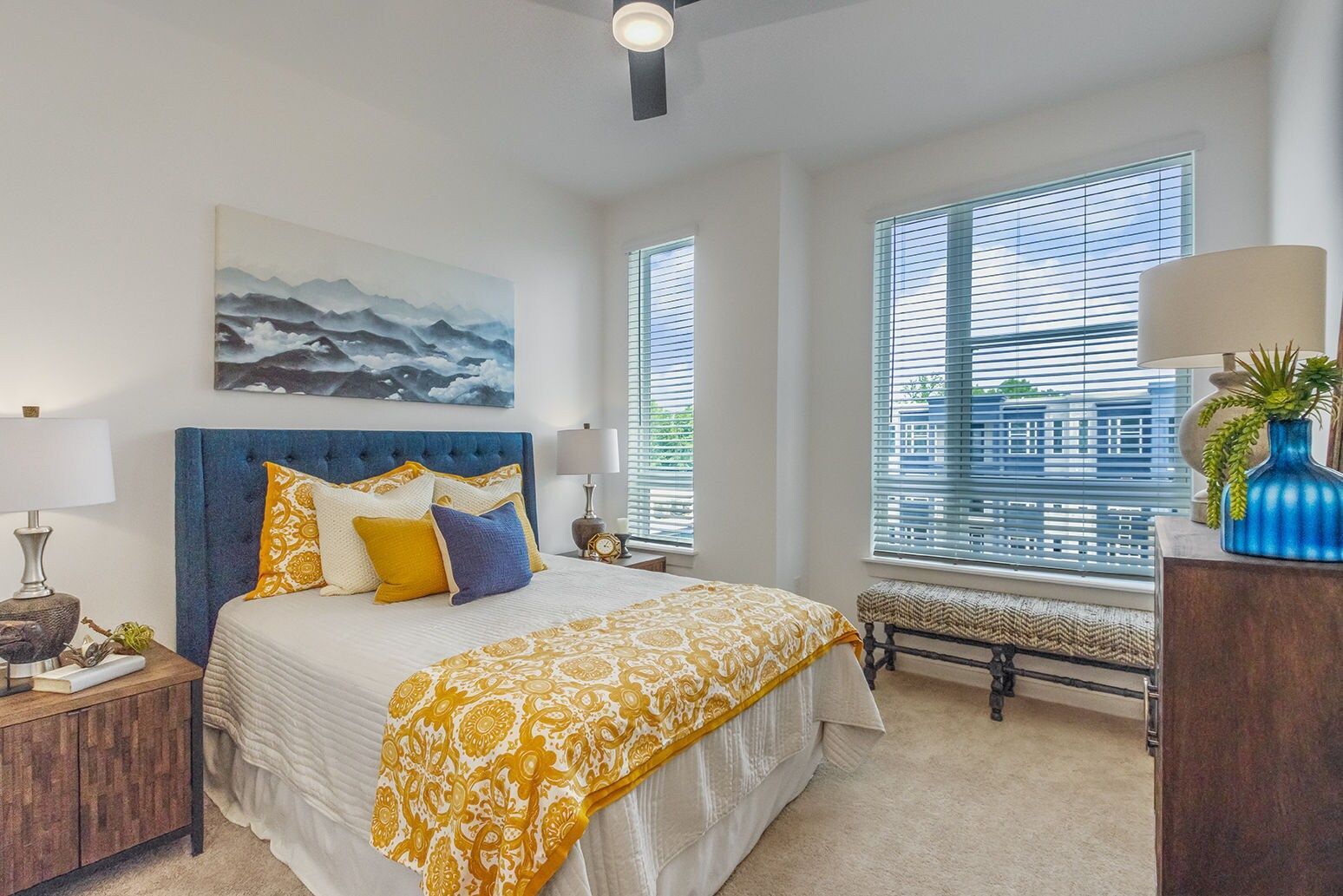 Bedroom with a blue tufted headboard, gold and blue bedding, and a large window with blinds.