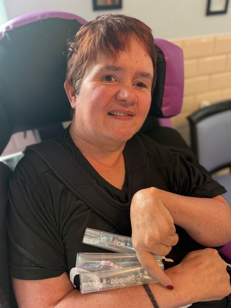 Woman with brown hair in a chair, pointing down at several items she holds. Smiling, indoor setting.