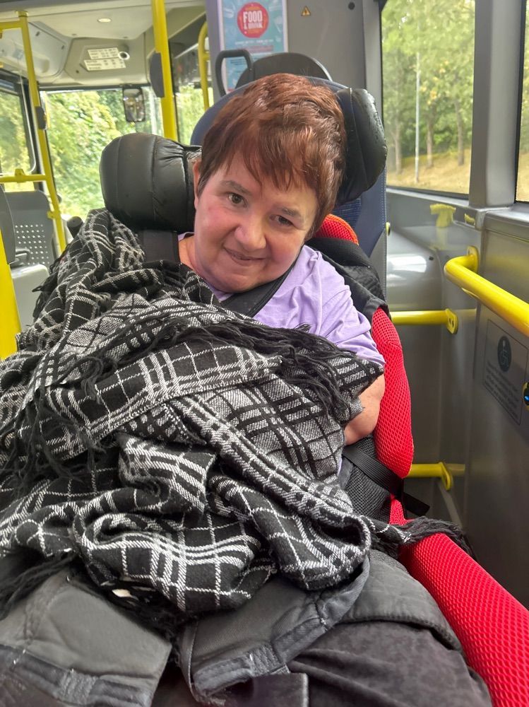 Woman in wheelchair on bus, wrapped in plaid blanket, smiling.
