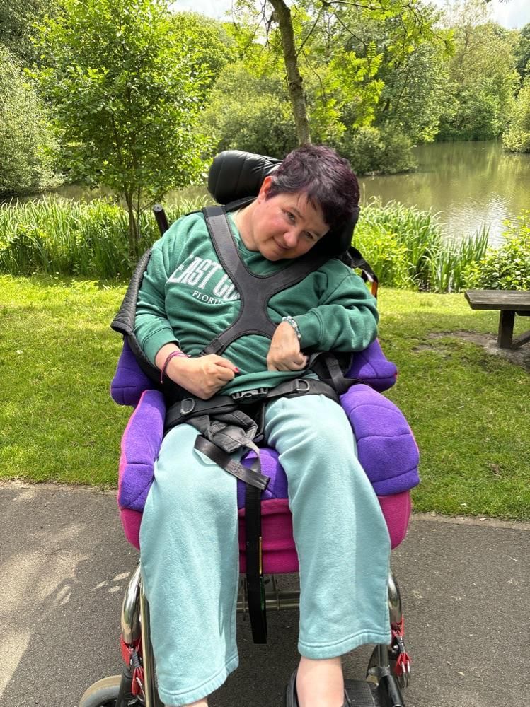 Woman in a wheelchair by a lake, wearing green sweatshirt and blue pants, smiling.