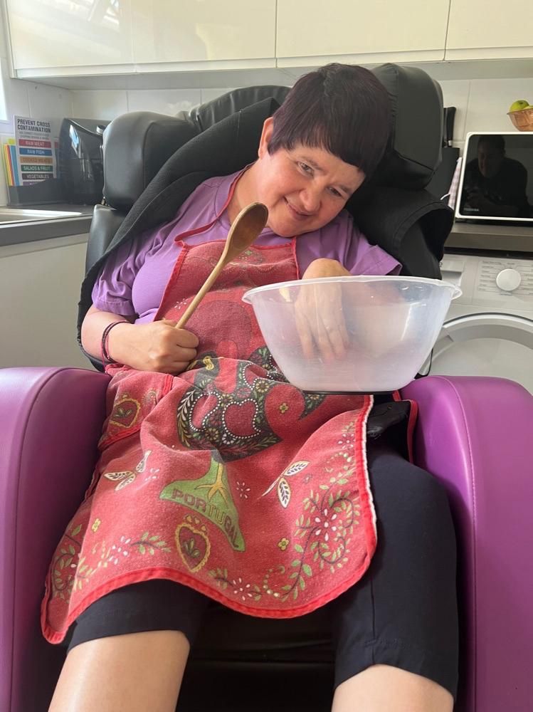 Woman in a wheelchair, wearing an apron, smiling while holding a wooden spoon and bowl in a kitchen.