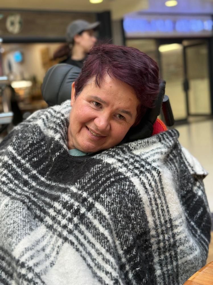 Woman in wheelchair wrapped in a black and white blanket, smiling, with short red hair, indoors.