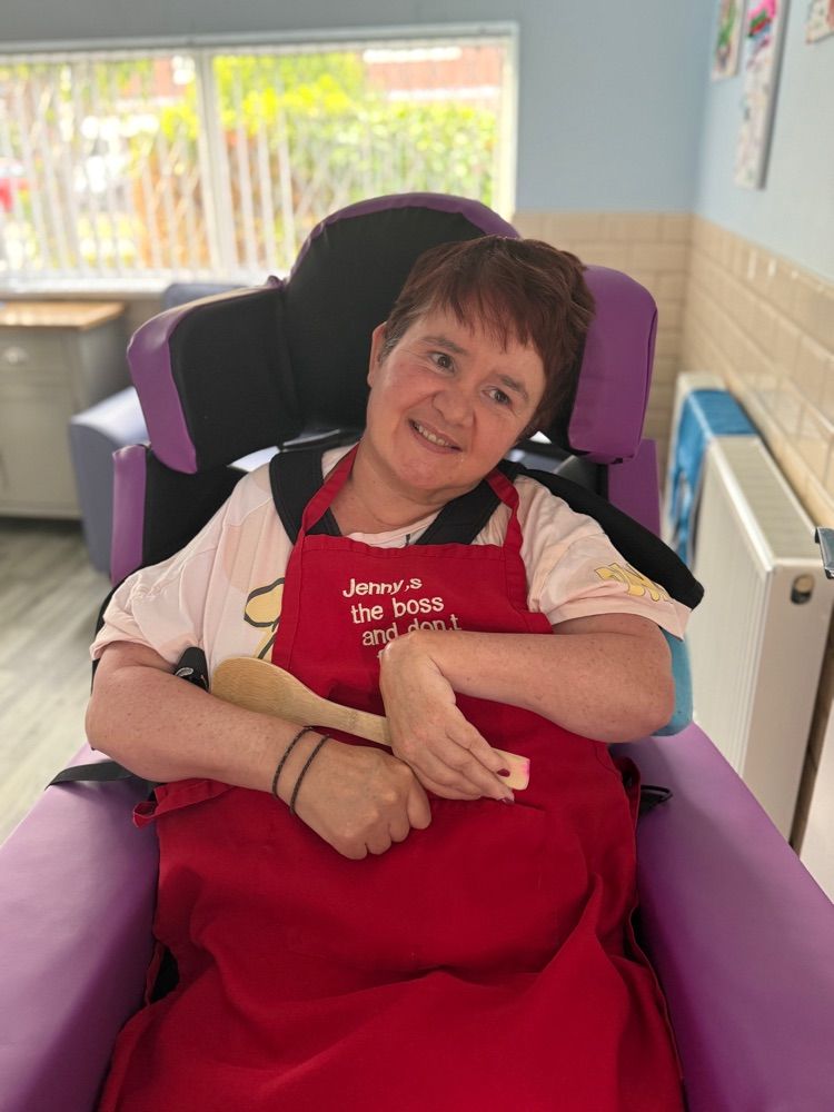 Woman in a wheelchair, wearing a red apron, smiles while holding a wooden spoon.