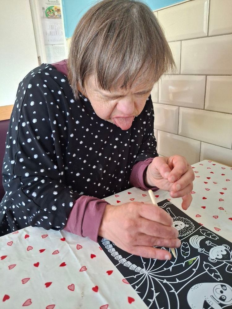 Woman with Down syndrome focused on drawing on black scratch art at a table.