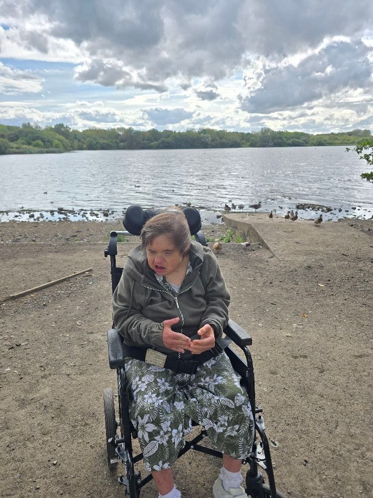 Woman in wheelchair by lake, wearing camouflage jacket and pants, cloudy sky.