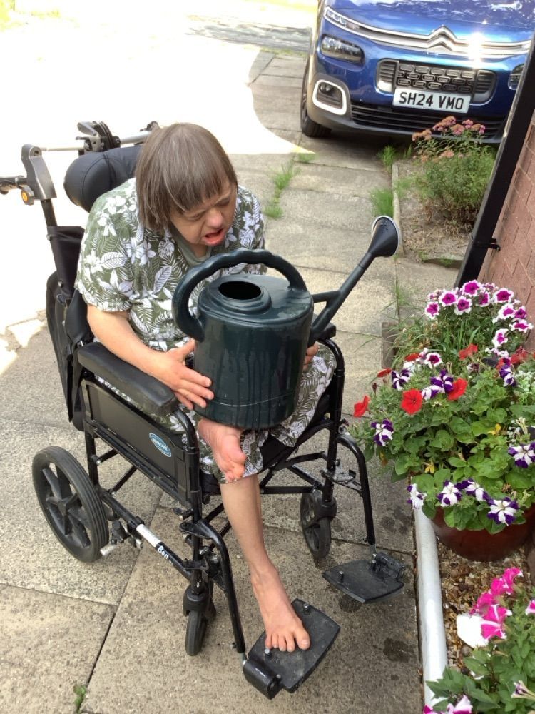 Woman in wheelchair watering flowers with a green watering can outside.