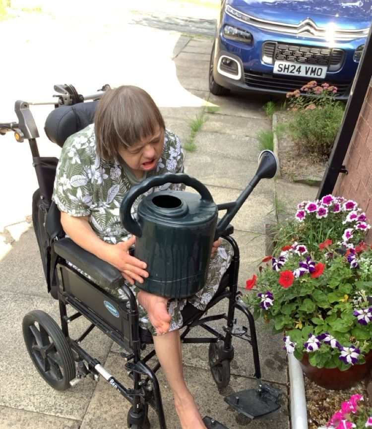 Person in wheelchair waters flowers with a green watering can outside. Blue car in background.